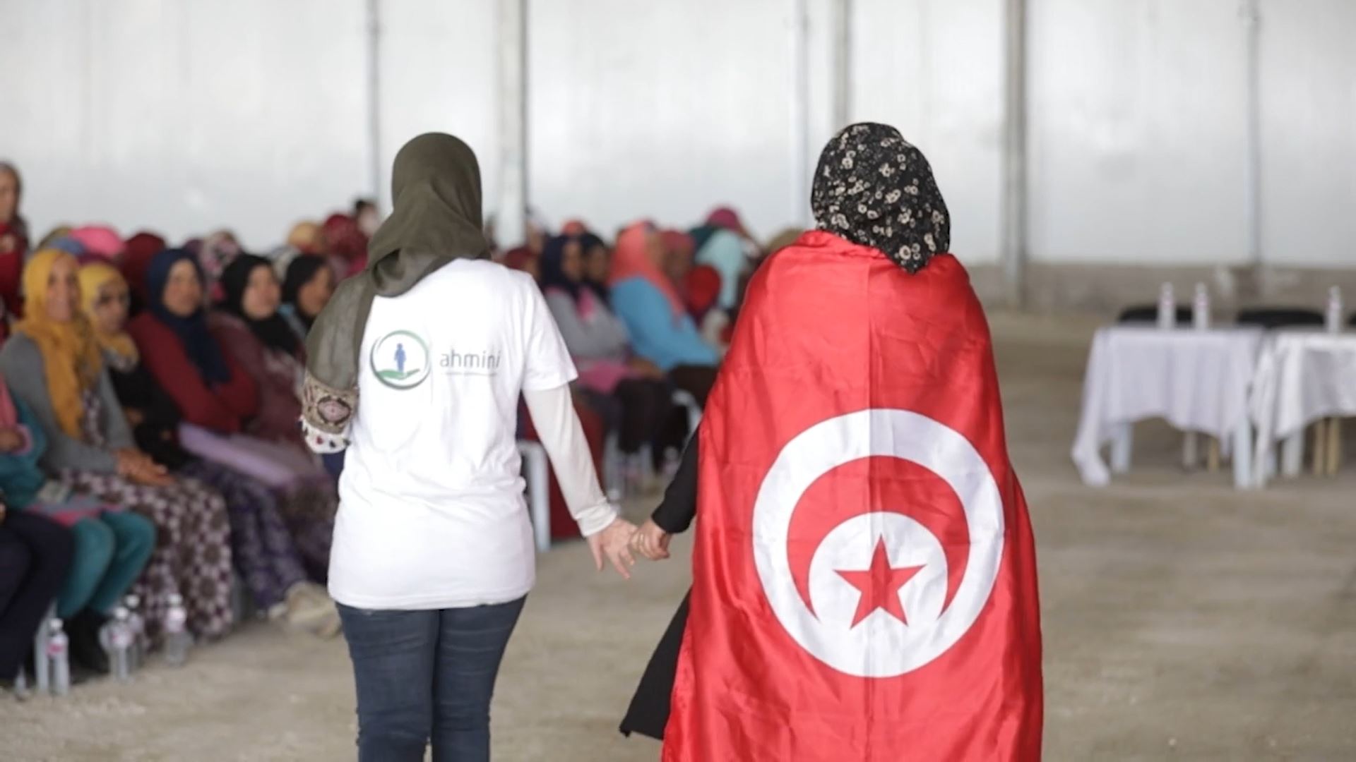 Backs of two women walking next to a seated crowd, one wearing a Tunisia flag